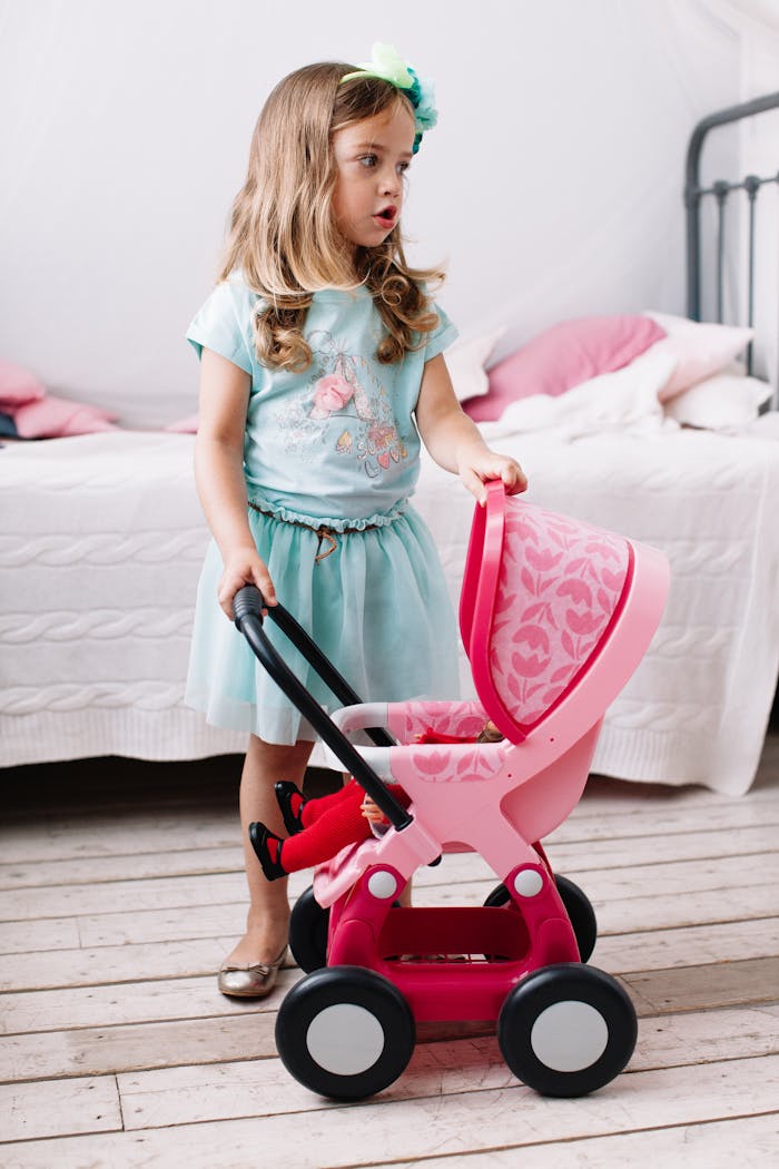 A young girl in a pastel dress playing indoors with a pink toy stroller.